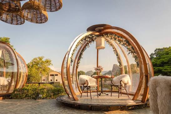 A transparent timber-ribbed dining dome on a deck, set with a round table, two sheepskin-draped chairs and a hanging lantern