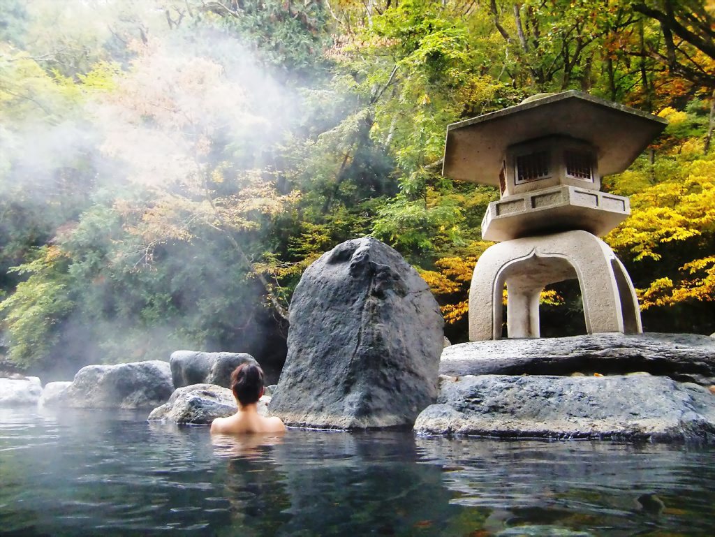 A bather in an open-air thermal rock pool below a stone lantern and autumn trees